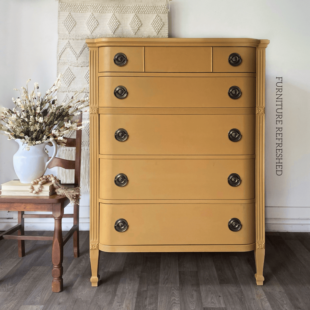 Photo of a vintage 5 drawer tallboy dresser. The dresser is painted yellow and has aged bronze Hepplewhite style hardware. A wooden chair with some books and a vase are beside the cresser.