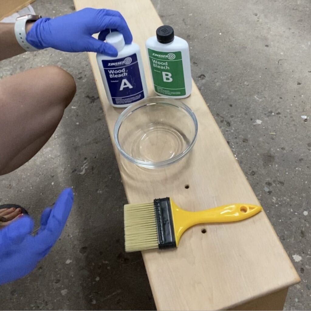 Image of a vintage dresser drawer standing upright with all of the stain removed. On the top is the Zinsser wood bleach two part product, along with a chip brush and a glass bowl. 