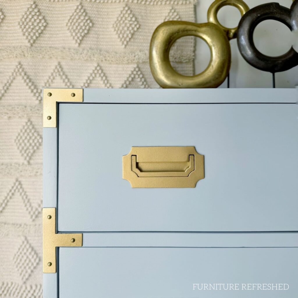 Close up view of the top corner of a blue campaign style painted dresser. Shows blue paint and gold hardware.