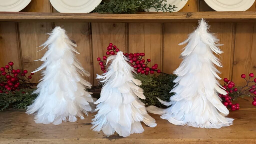 Set of three feather Christmas trees on a shelf with evergreen branches and red berries behind them.