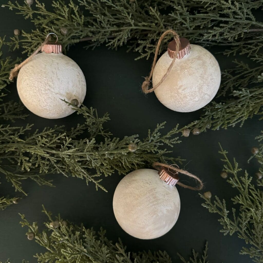 Three textured Christmas ornaments displayed on a green table iwth greenery surrounding them,