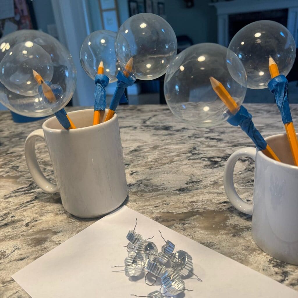 Glass ornaments that are ready for paint. Metal toppers have been removed and a pencil has been taped to the inside. The are resting upside down on the pencils in mugs so that they won't touch each other when drying.